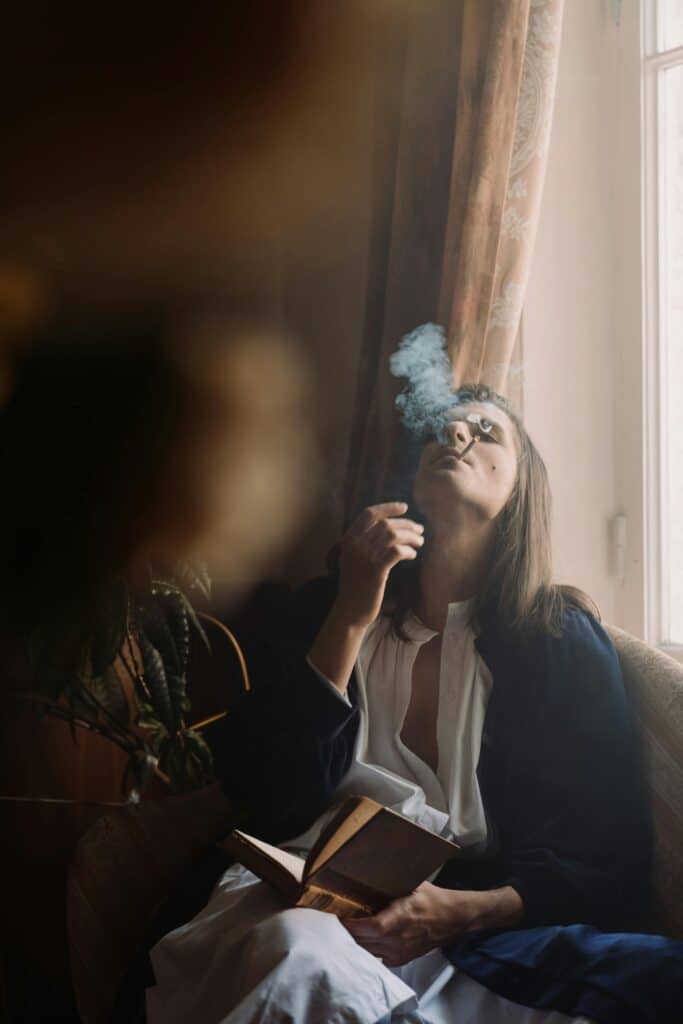 Person sitting indoors near a window, holding a book while exhaling smoke upward.