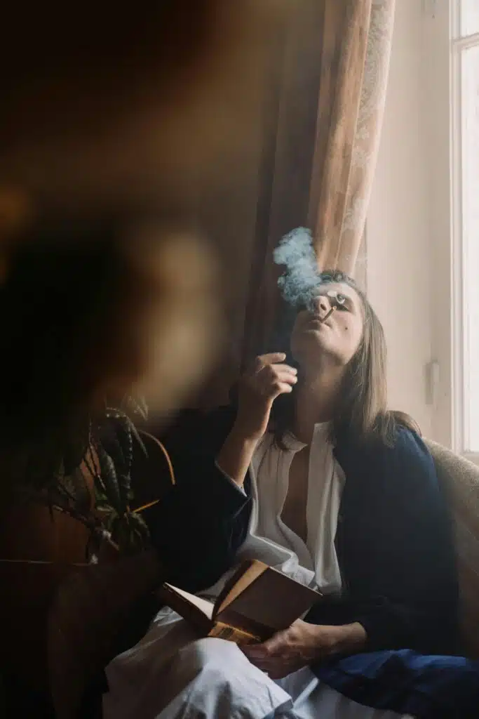 Person sitting indoors near a window, holding a book while exhaling smoke upward.