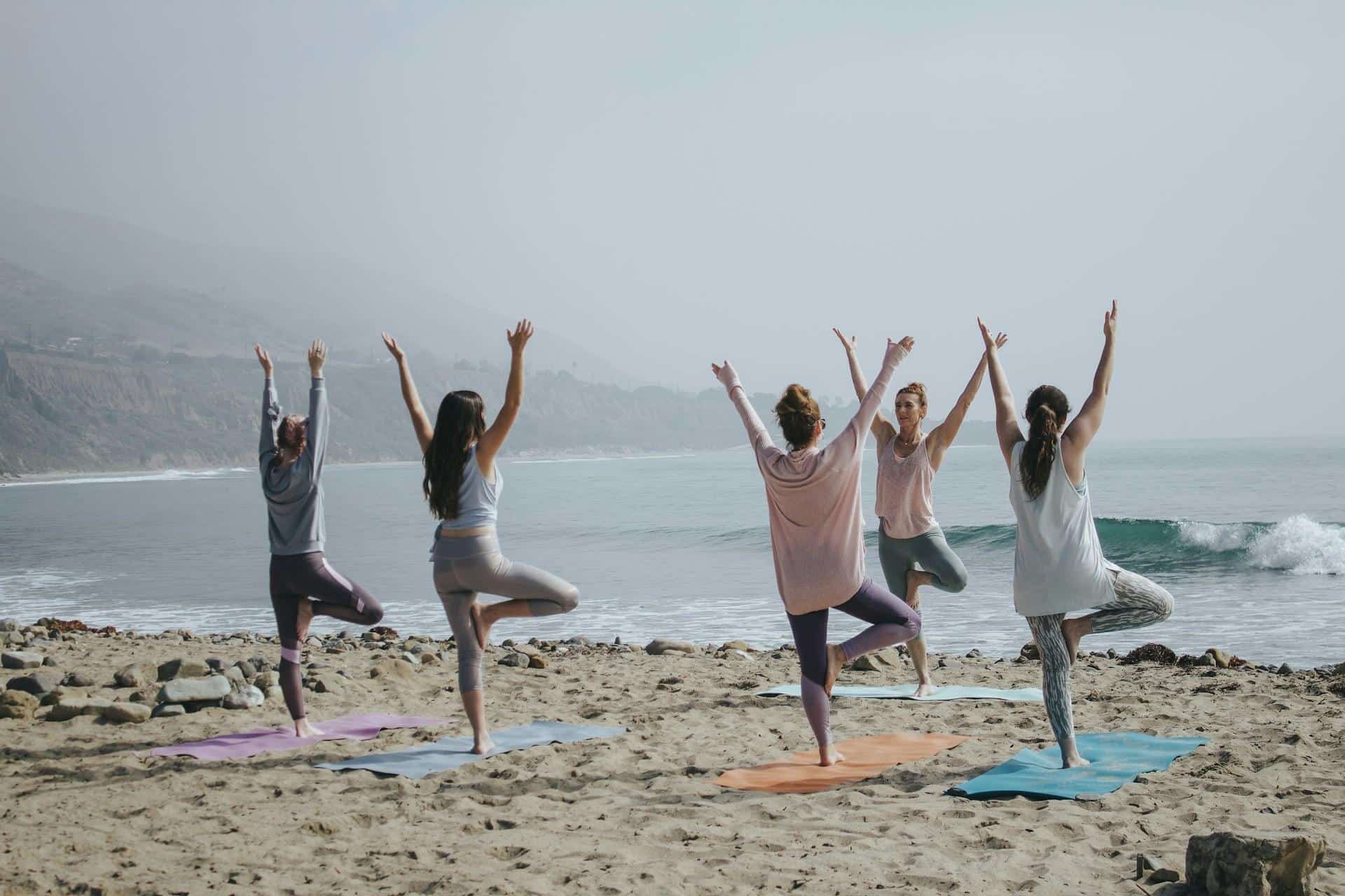Group of five women practicing tree pose yoga on colorful mats on a foggy beach, representing sober wellness activities and community events